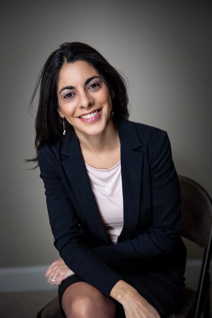A woman in a suit is sitting in a chair with her arms crossed and smiling.
