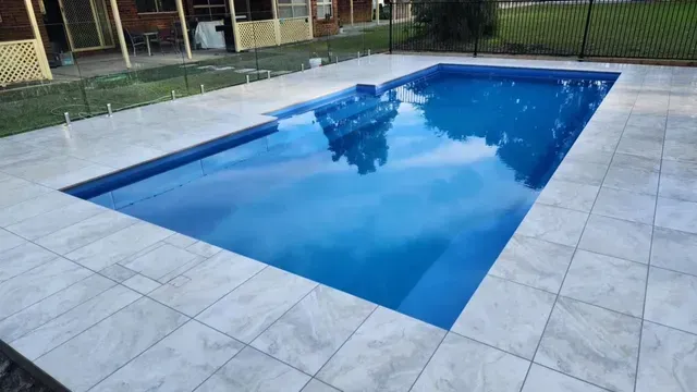 Rectangular backyard swimming pool with blue water beside a light stone patio, reflecting trees and sky