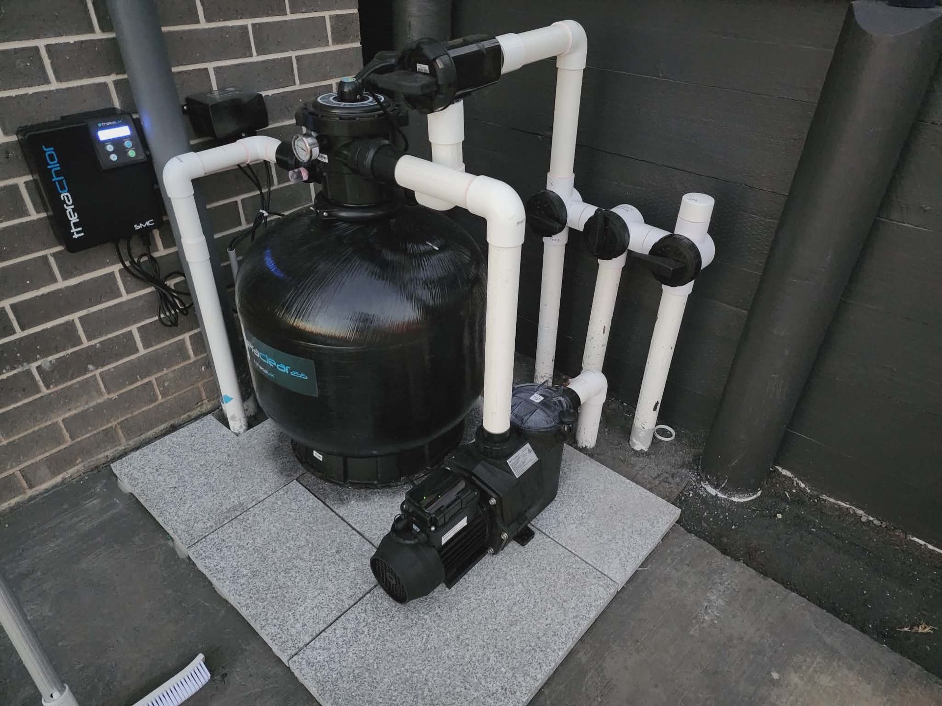 Pool equipment: filter, pump, pipes on a gray tile base, against a brick wall.  — Clearwater Pools Hallidays Point in Hallidays Point, NSW