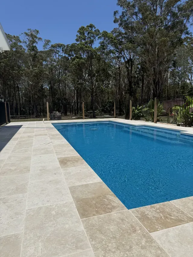 Rectangular blue swimming pool beside a light stone deck, with trees lining the background.