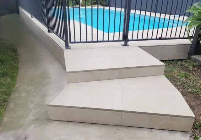 Tiled steps beside a fenced swimming pool, with a concrete path and grass nearby