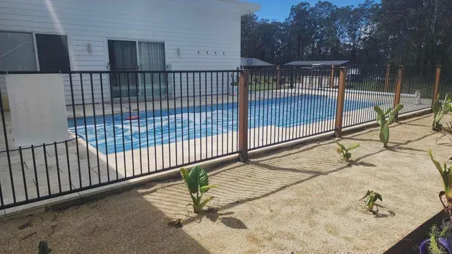 Backyard pool enclosed by black metal fence, with tan patio and potted plants in the foreground