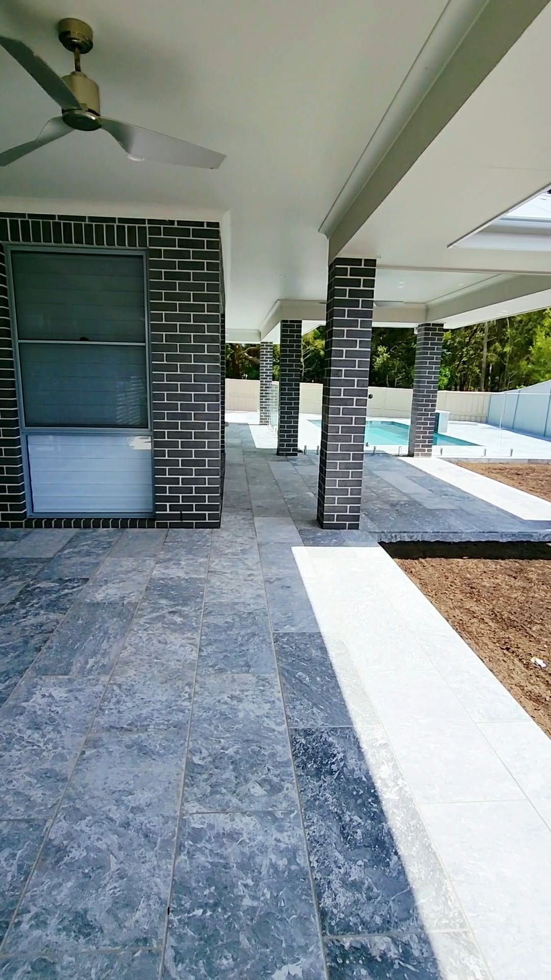 Covered Outdoor Patio With Dark Brick Pillars, Tiled Floor, and a Swimming Pool Visible in the Distance — Clearwater Pools Hallidays Point in Hallidays Point, NSW