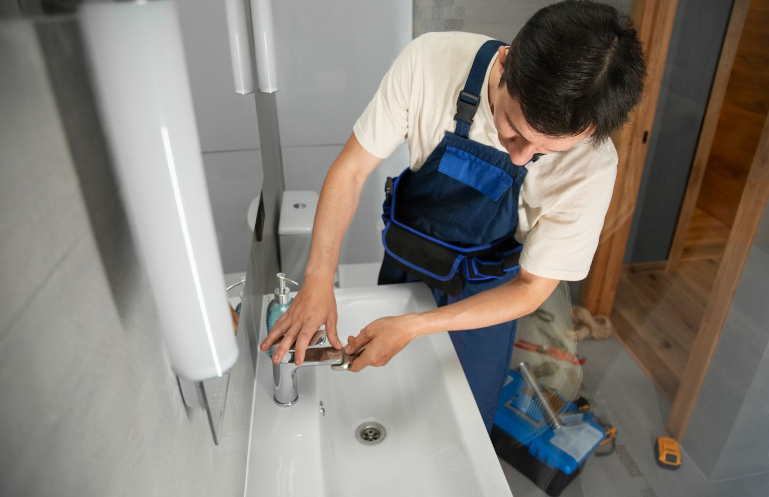Plumber in blue overalls repairing a sink in a bathroom.