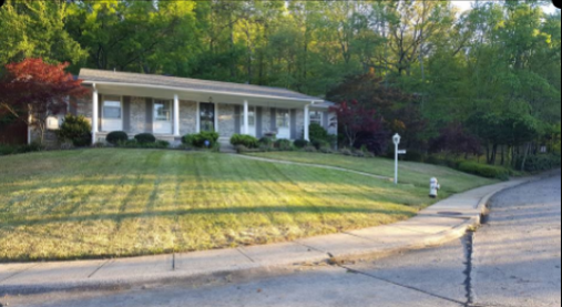 A single-story house with a green lawn. Trees in the background, a sidewalk and driveway in the foreground.