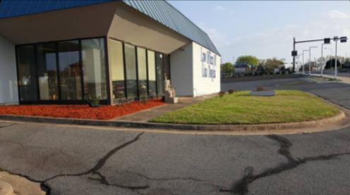Exterior of a small white building with large windows, red mulch, green grass, and cracked asphalt.