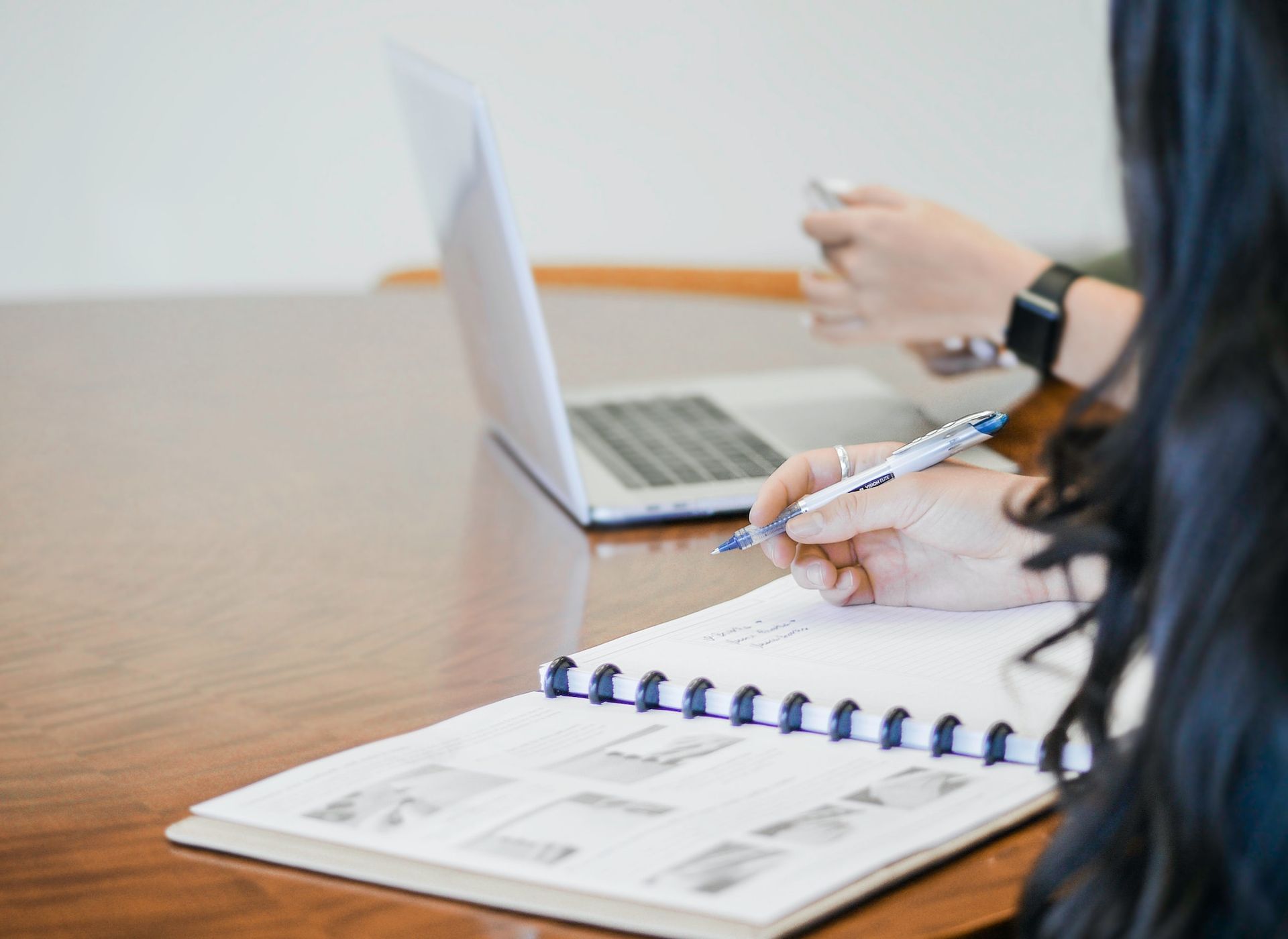 A woman is writing in a notebook in front of a laptop computer.