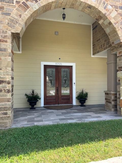 Exterior view of a home with arched stone entrance, wooden double doors, and potted plants. Yellow siding and gray paving.
