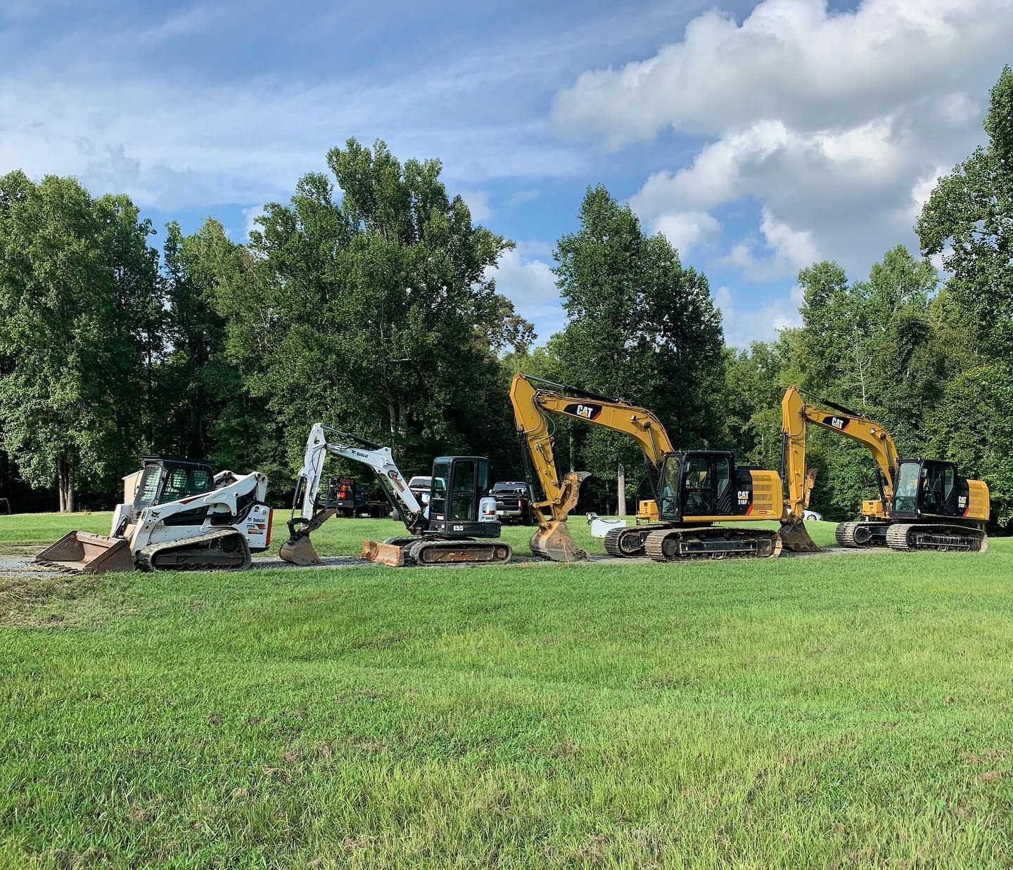 Lineup of construction excavators on green grass under a partly cloudy sky.