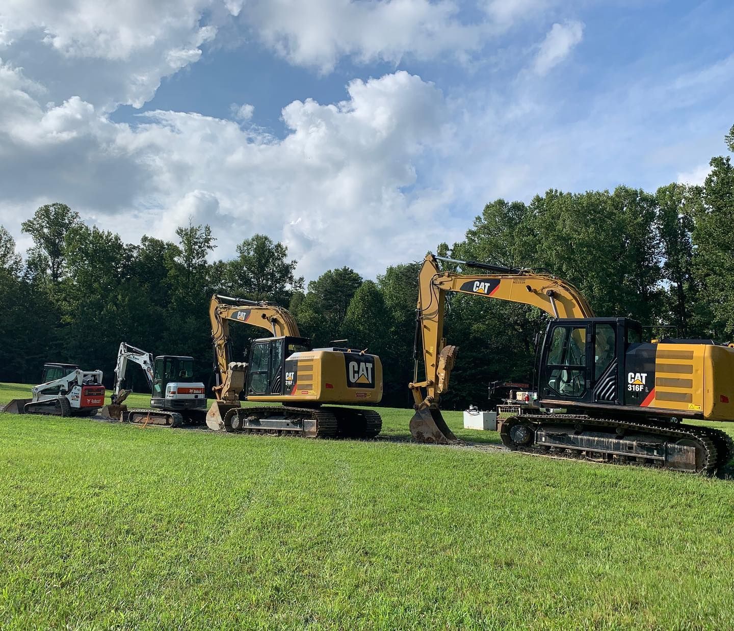 Several construction vehicles lined up in a grassy field. Yellow Caterpillar excavators, a Bobcat, and a ditch are visible.