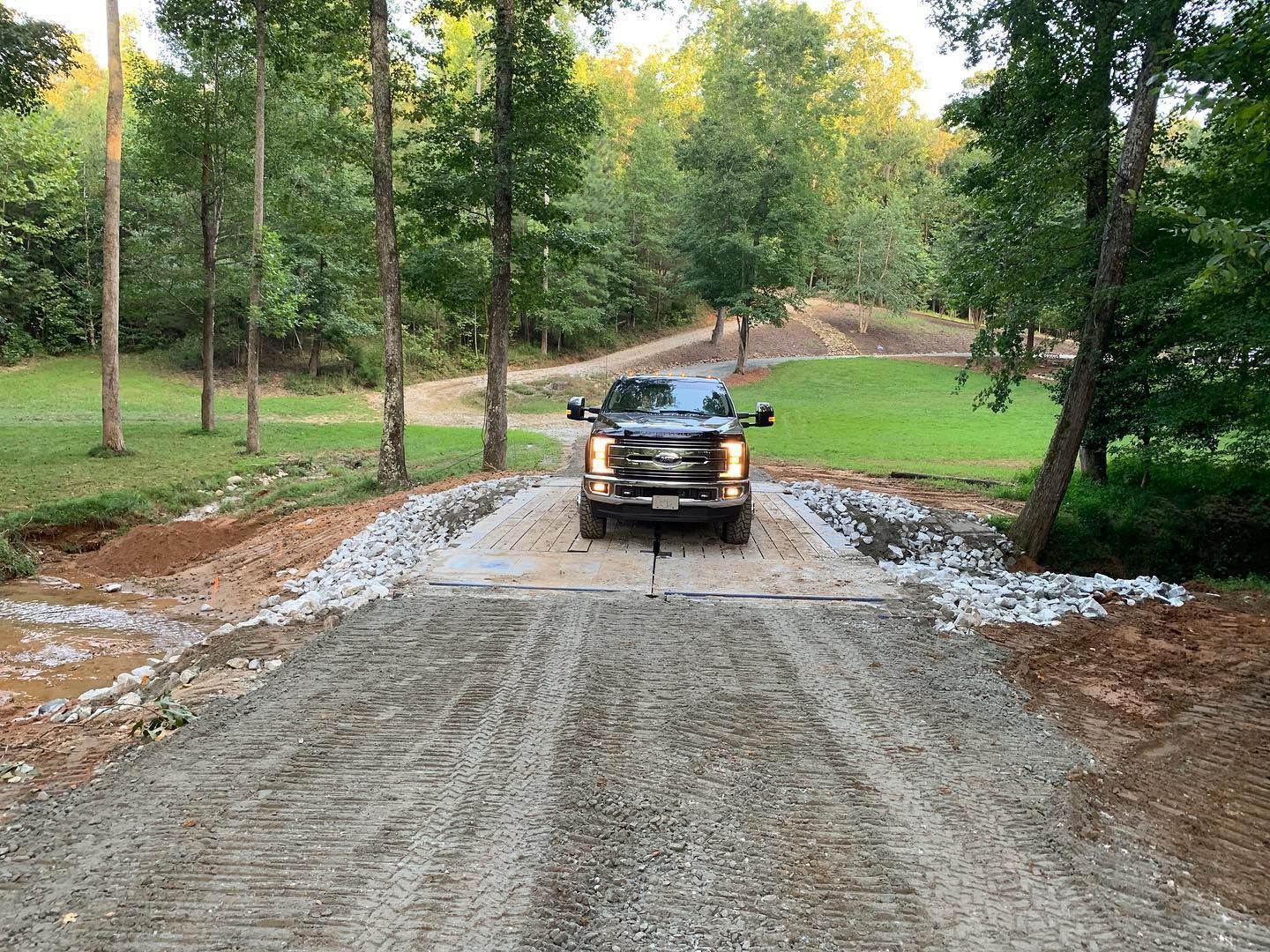 Truck driving over a gravel road crossing a creek, trees in the background.
