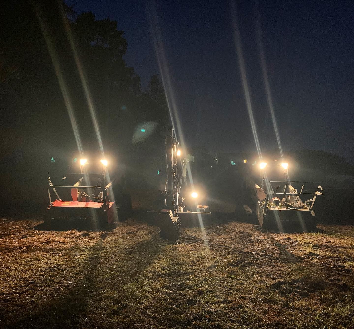Three tractors with bright headlights operate in a dark field at night.