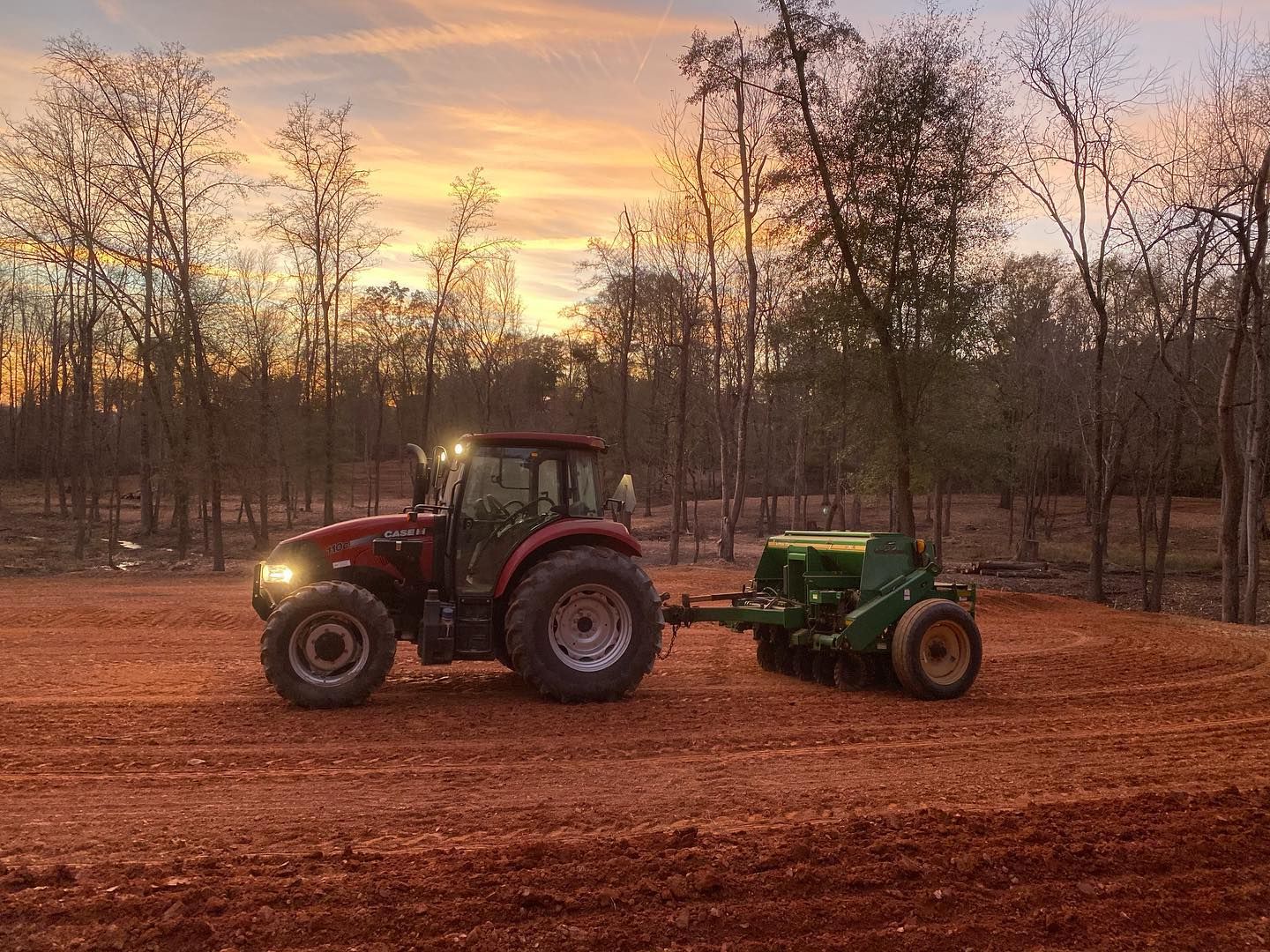 Tractor pulling a seeder across a field at sunset. Red tractor with green seeder, golden sky.
