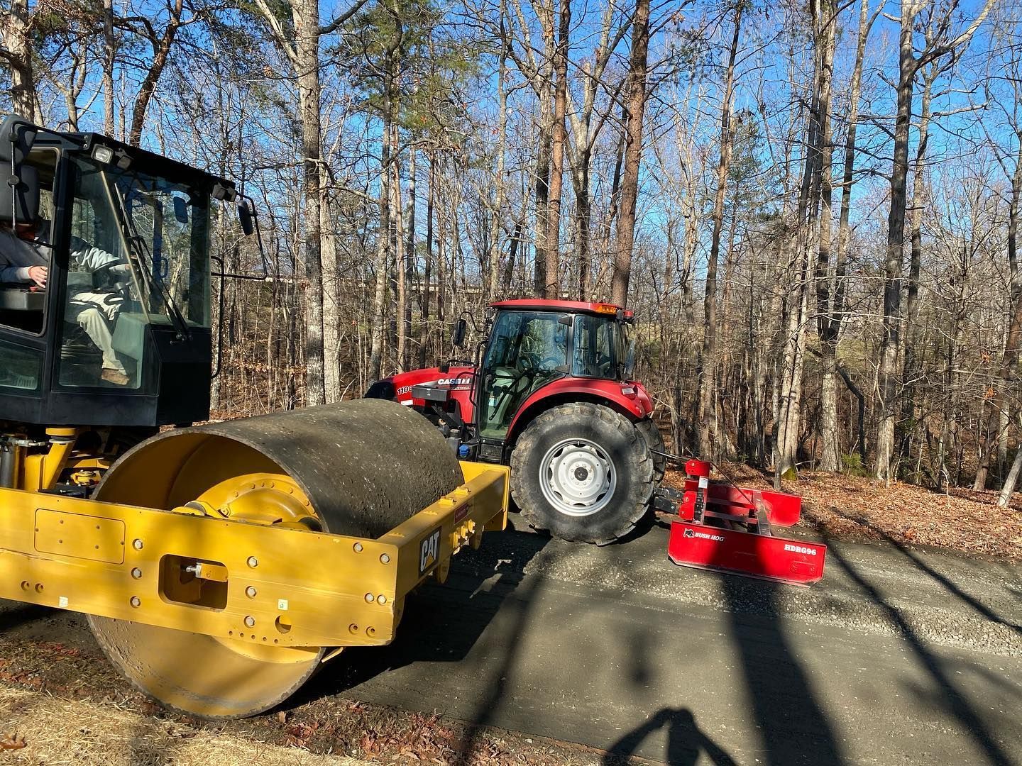 A road roller and red tractor compact a new asphalt road in a wooded area.