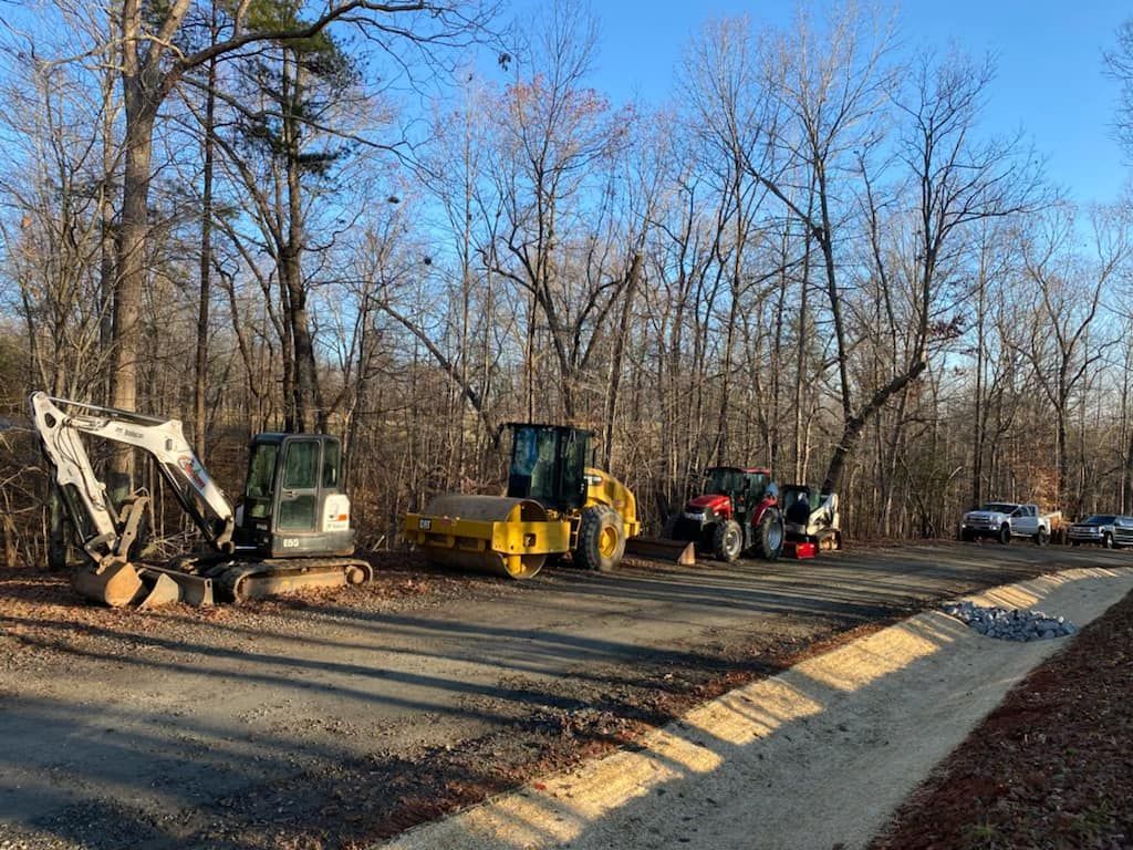 Construction site with heavy machinery like a backhoe, roller, and tractor on a gravel road, trees in background.