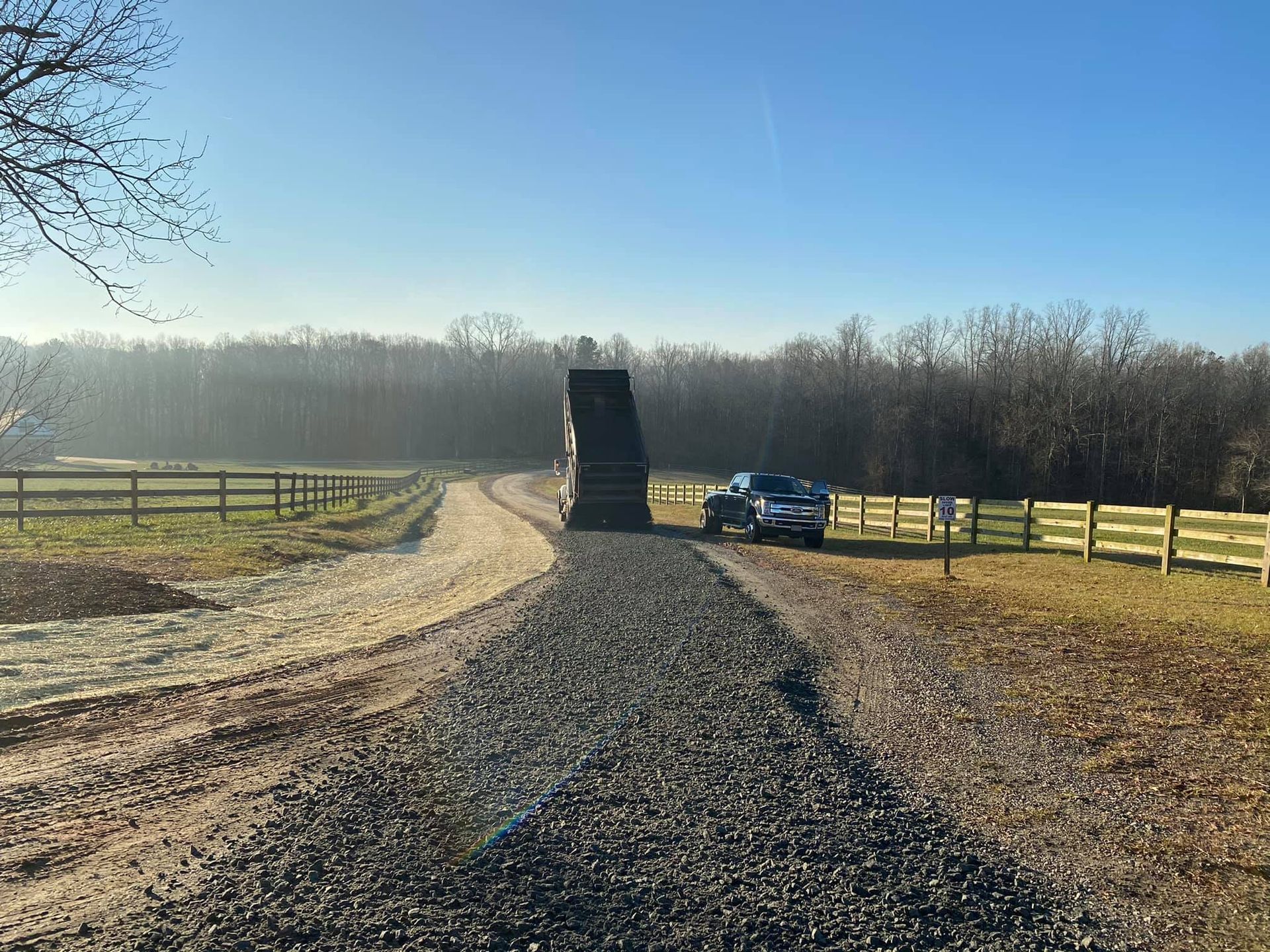Gravel driveway leading to a large object, with a truck parked to the side; fence and trees in background.
