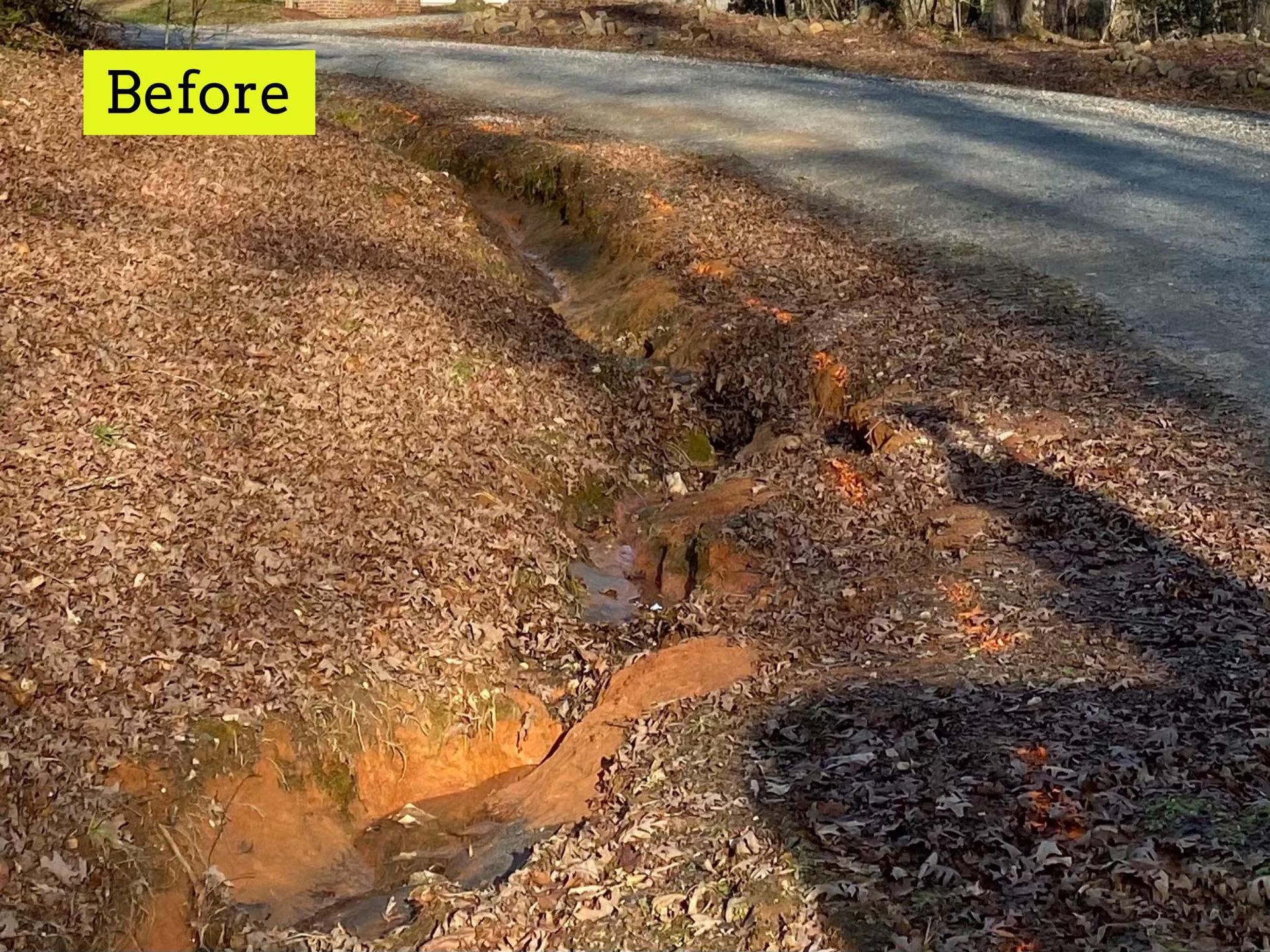 Erosion ditch beside a gravel road, before repair. Brown earth, leaves, and exposed orange soil.