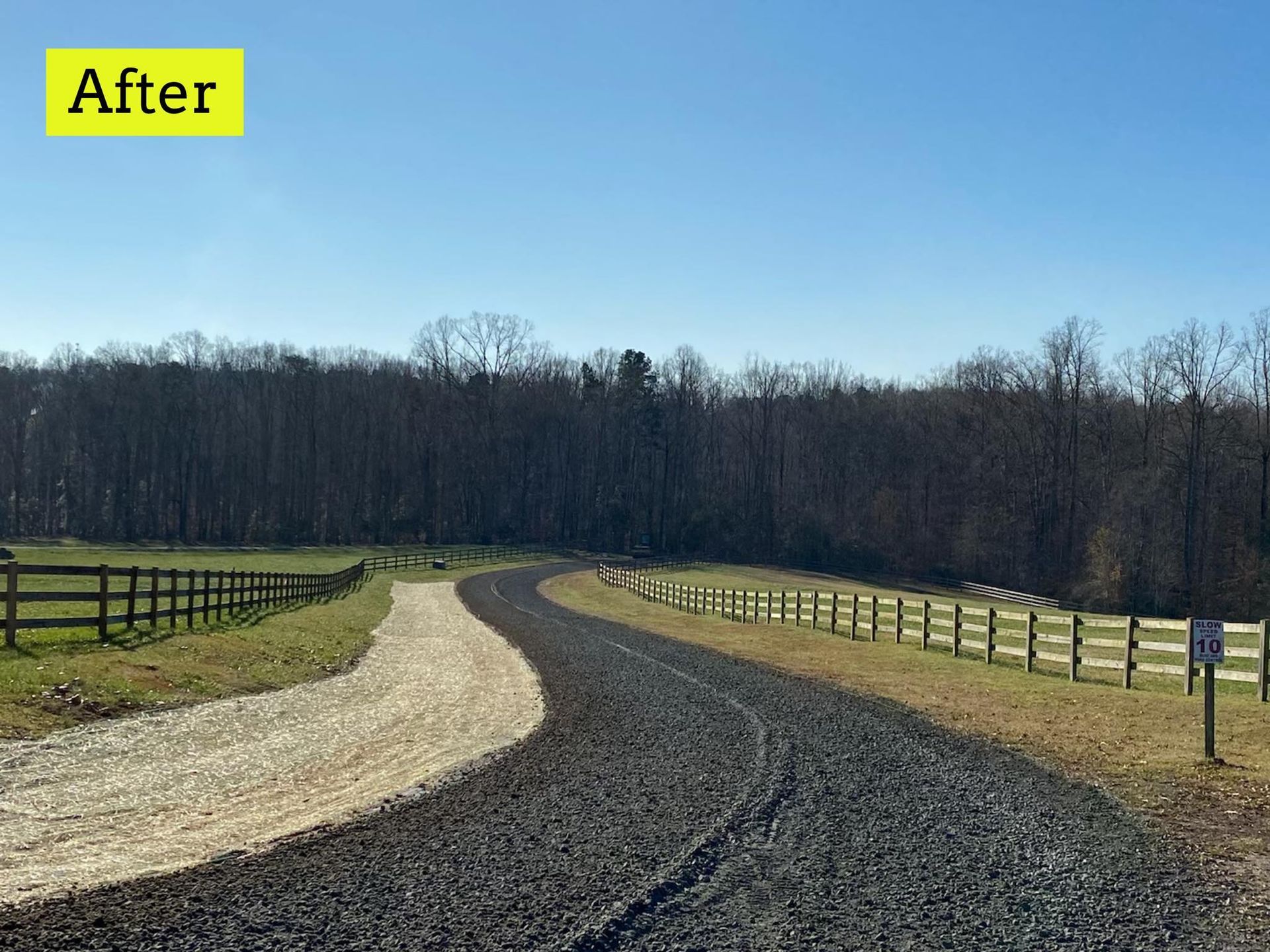 Gravel driveway with two sections, a light and dark color. Wooden fence borders green fields and trees.