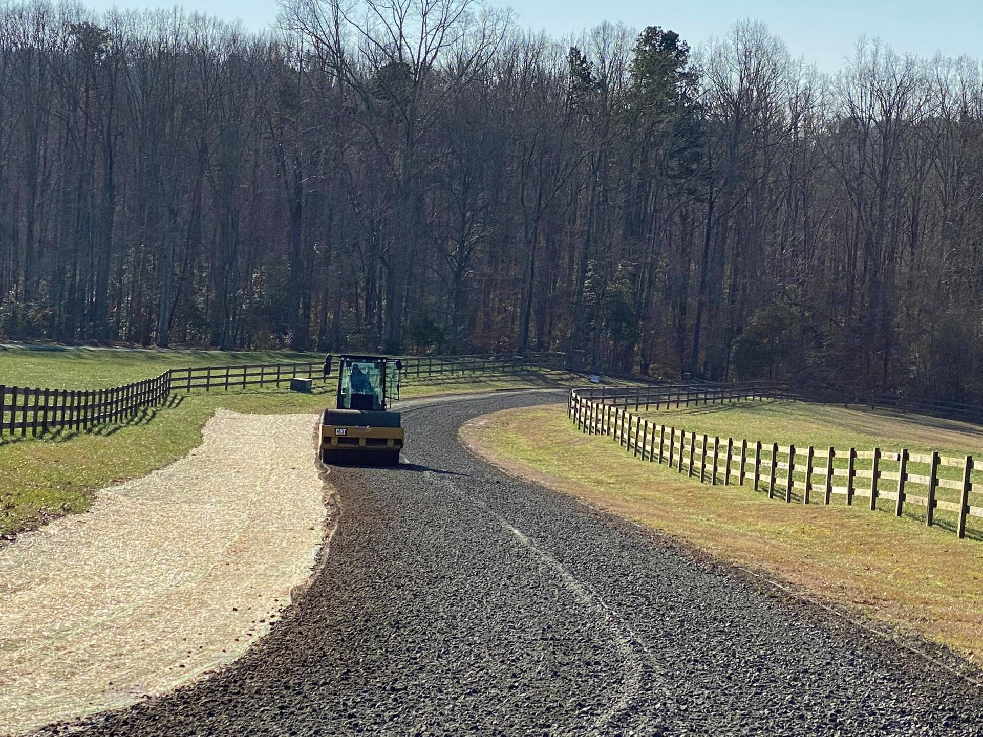 A roller compacting asphalt on a rural driveway. A new driveway side lies next to a fence and the trees in the background.