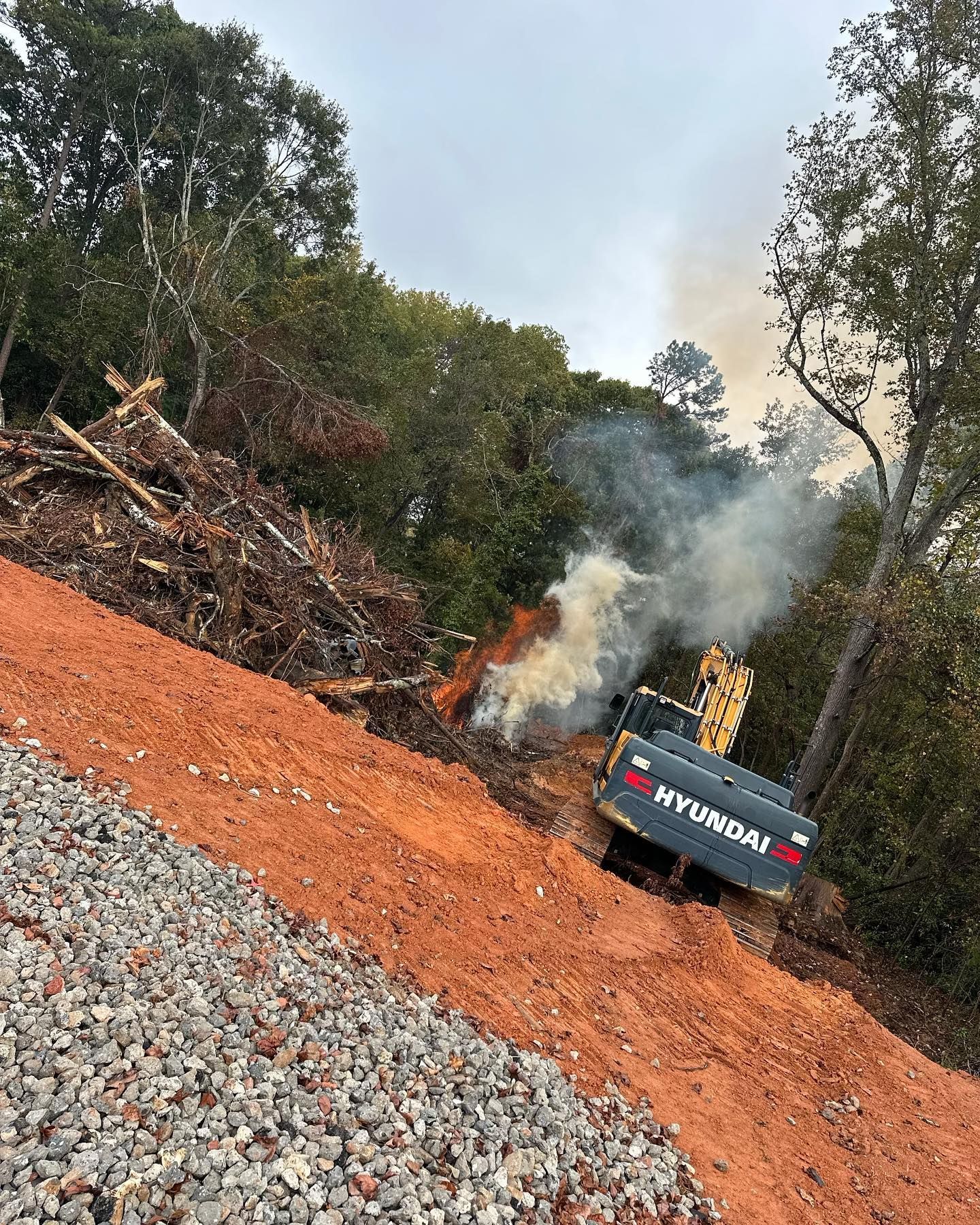 Hyundai excavator on a steep, red clay hillside, burning debris. Smoke rises near trees on an overcast day.