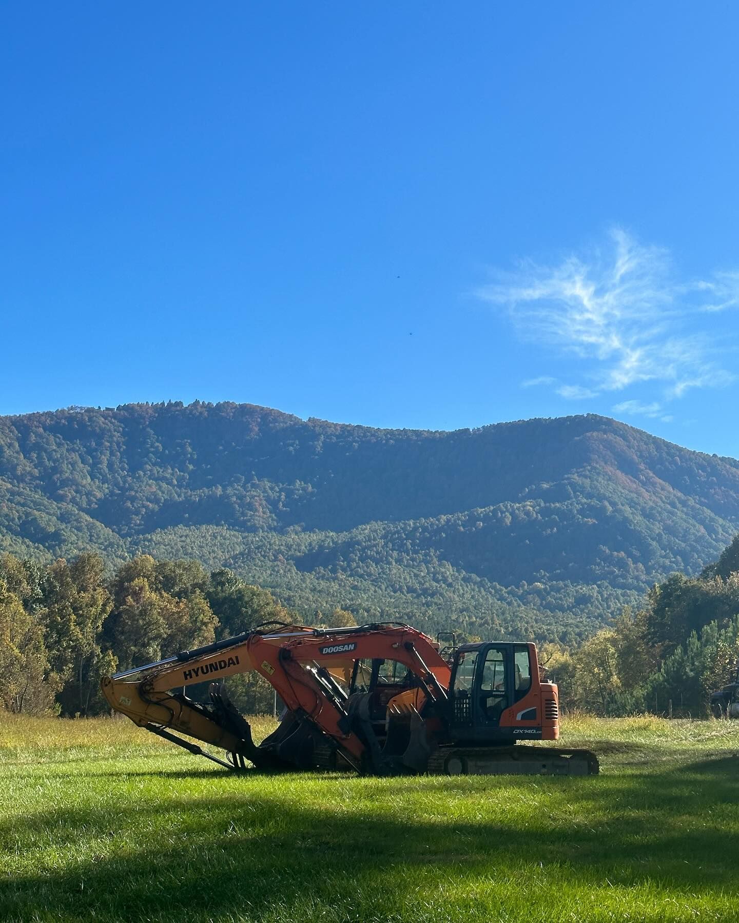 Orange excavator parked in a grassy field with a mountain backdrop under a blue sky.