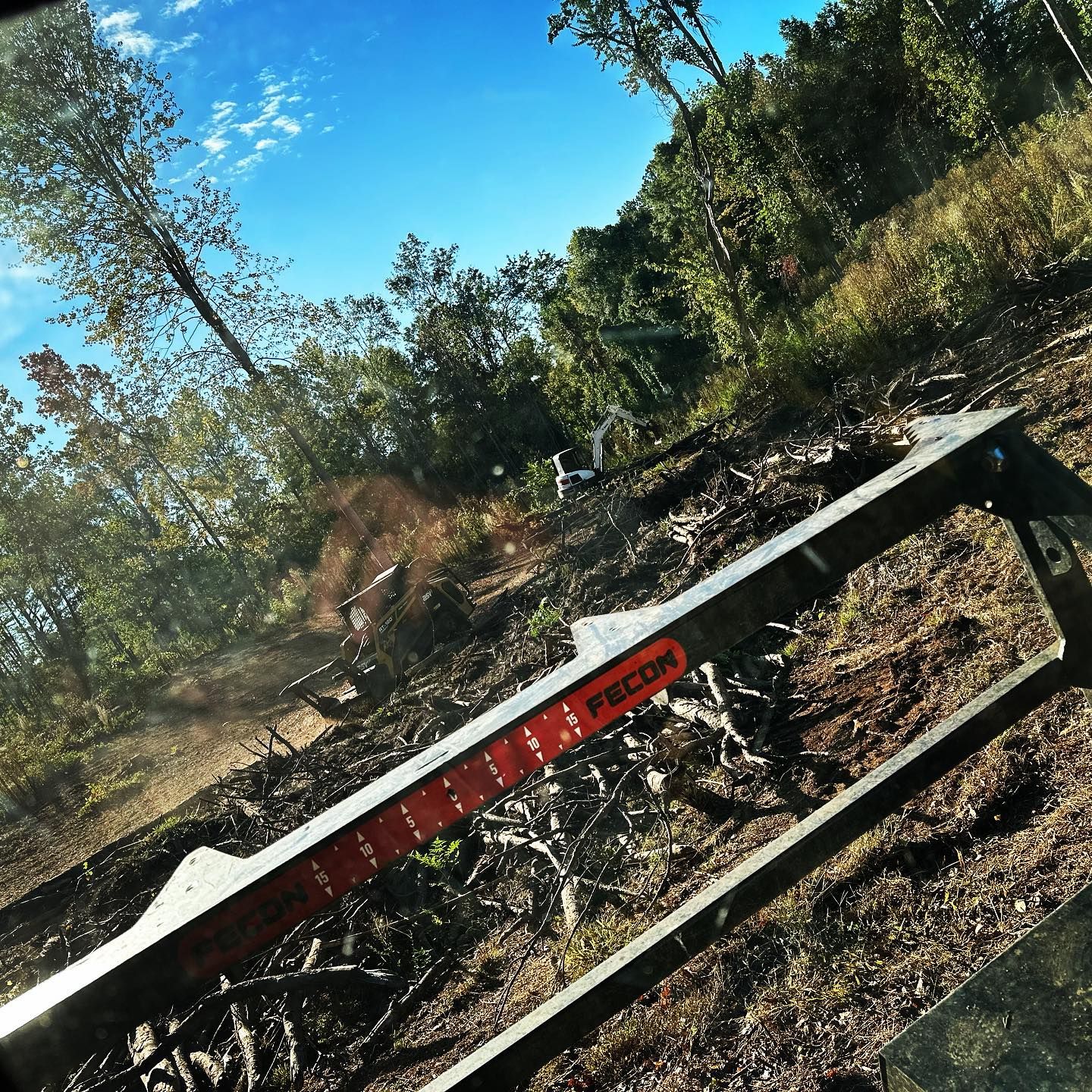 View from a vehicle window of recently cleared land with trees in background, bright sky.
