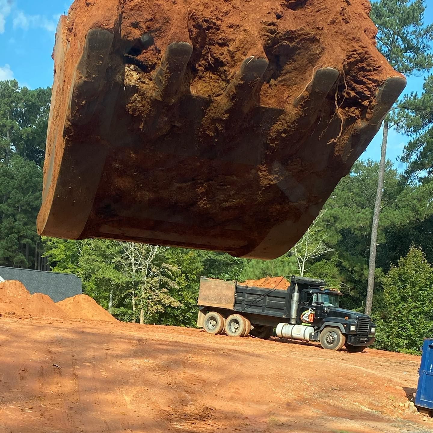 Excavator dumping red soil into a black dump truck at a construction site. Trees in the background.