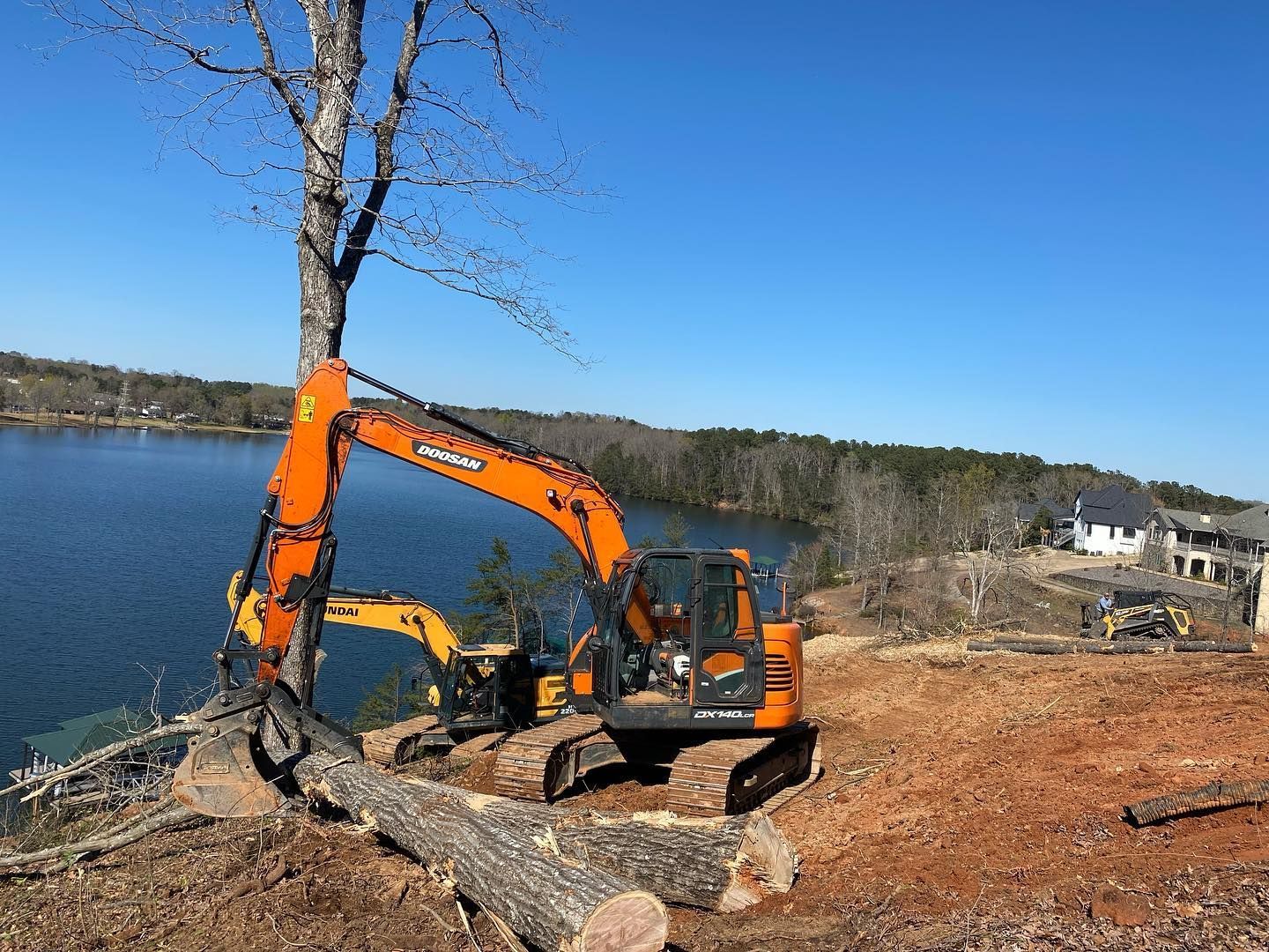 An orange excavator cutting a tree near a lake, sunny day, blue sky.