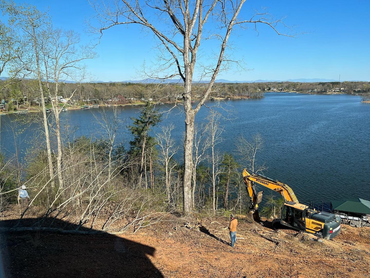 Excavator on a hillside overlooking a lake; bare trees and blue sky.