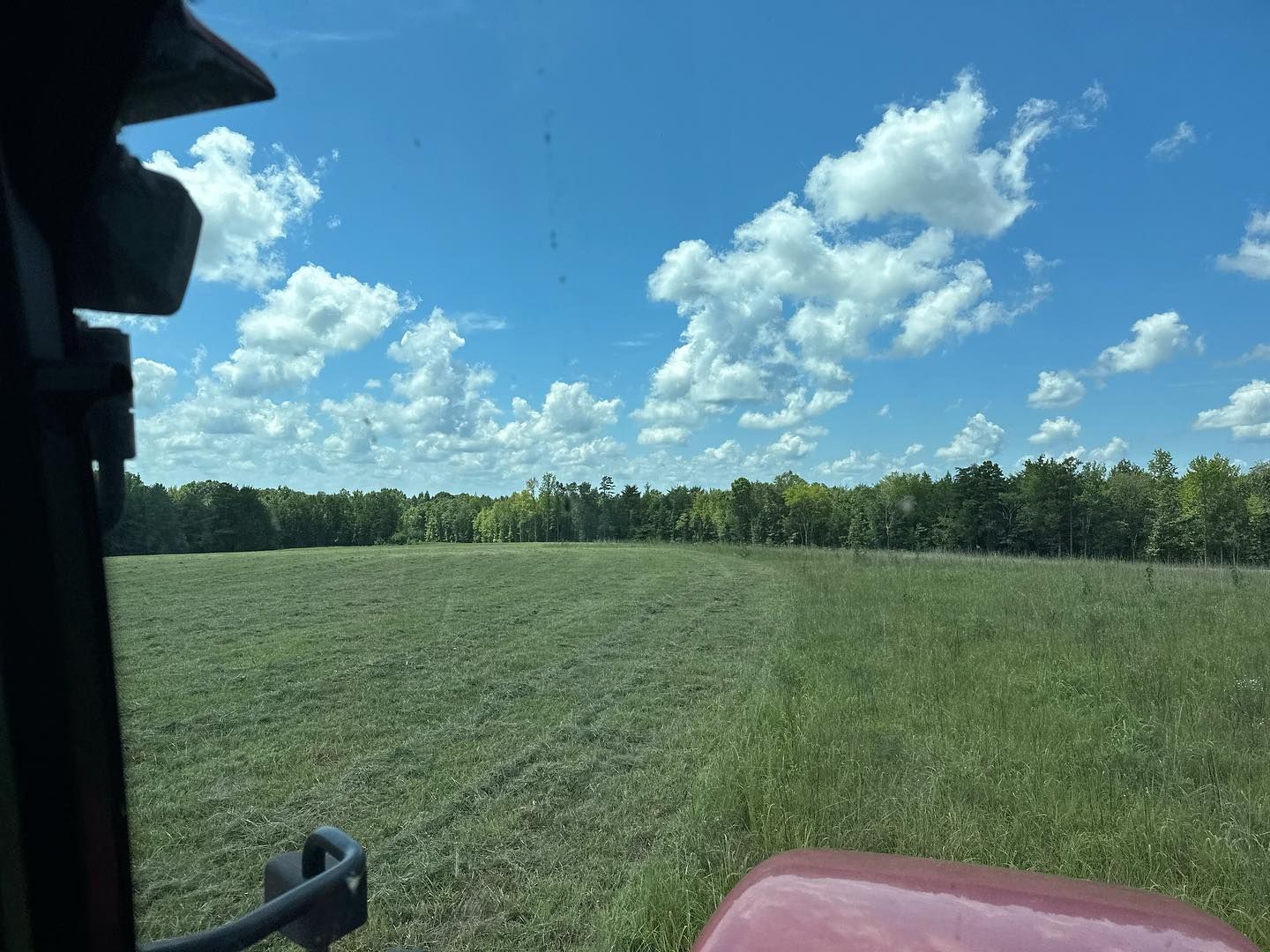 View from a tractor cab of a green field with trees under a blue sky with fluffy white clouds.