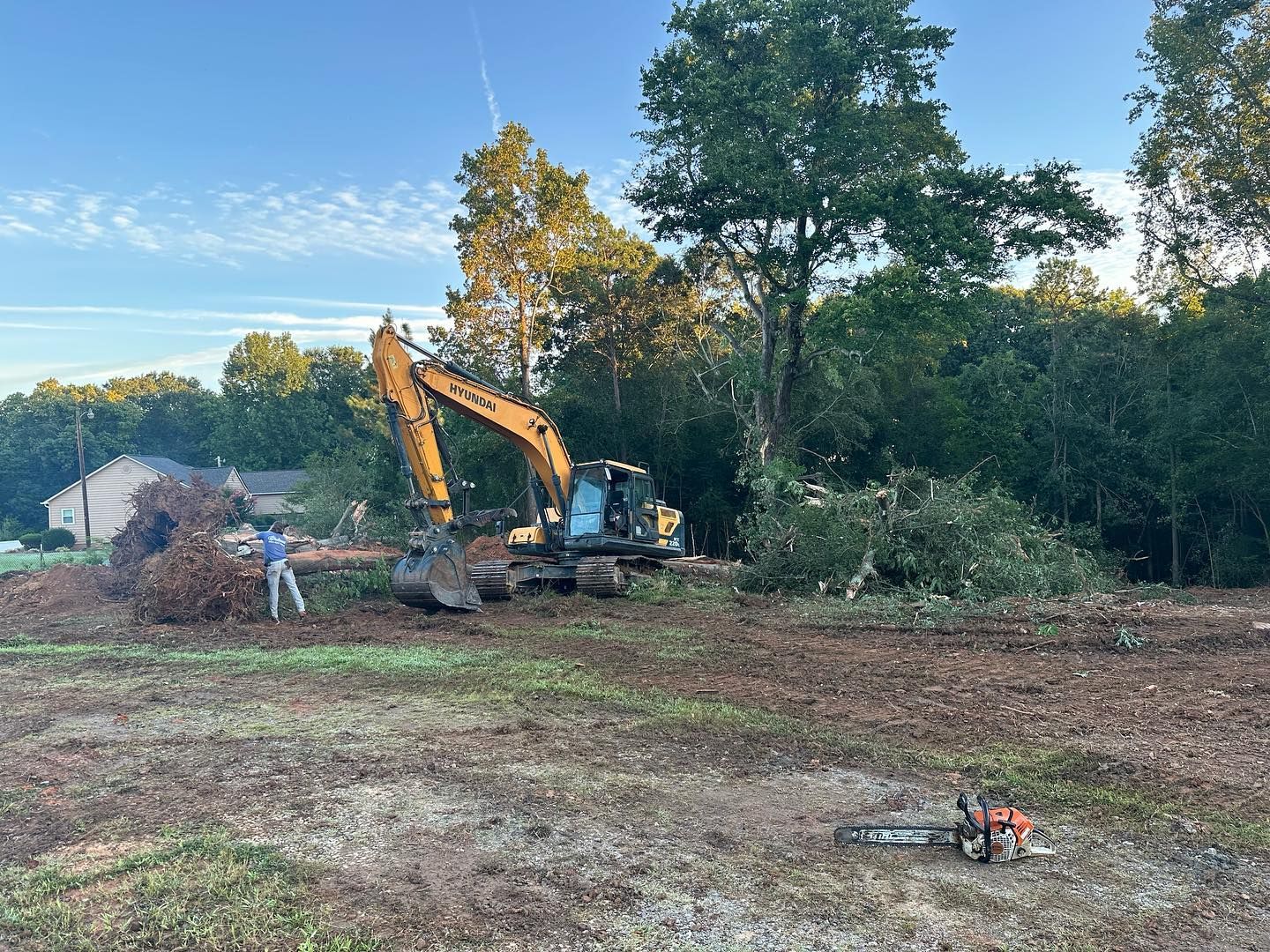 An excavator and a person are clearing land of brush. A chainsaw lies on the ground, trees in the background.