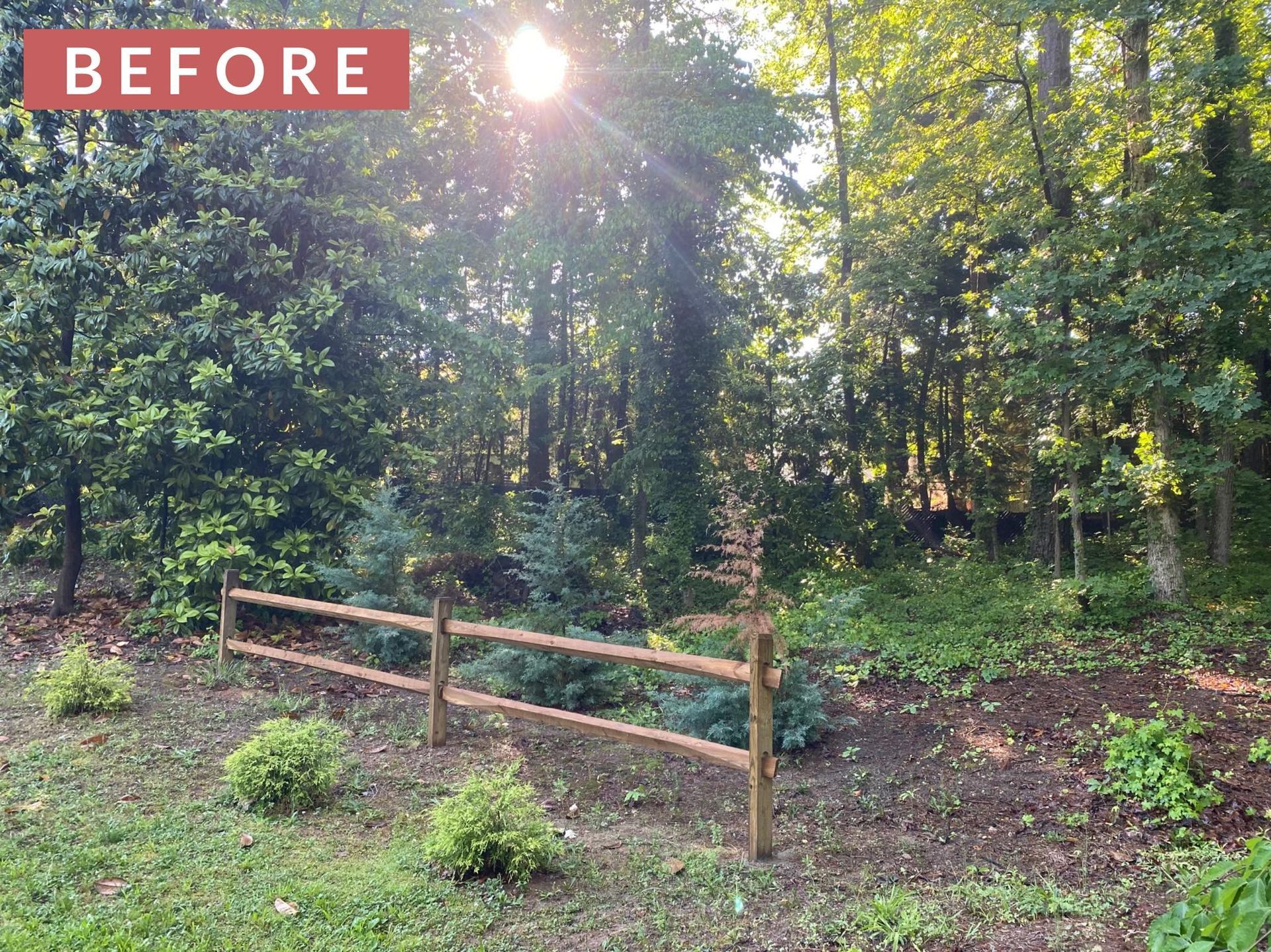 A rustic wooden fence in a wooded area with sun shining through the trees.