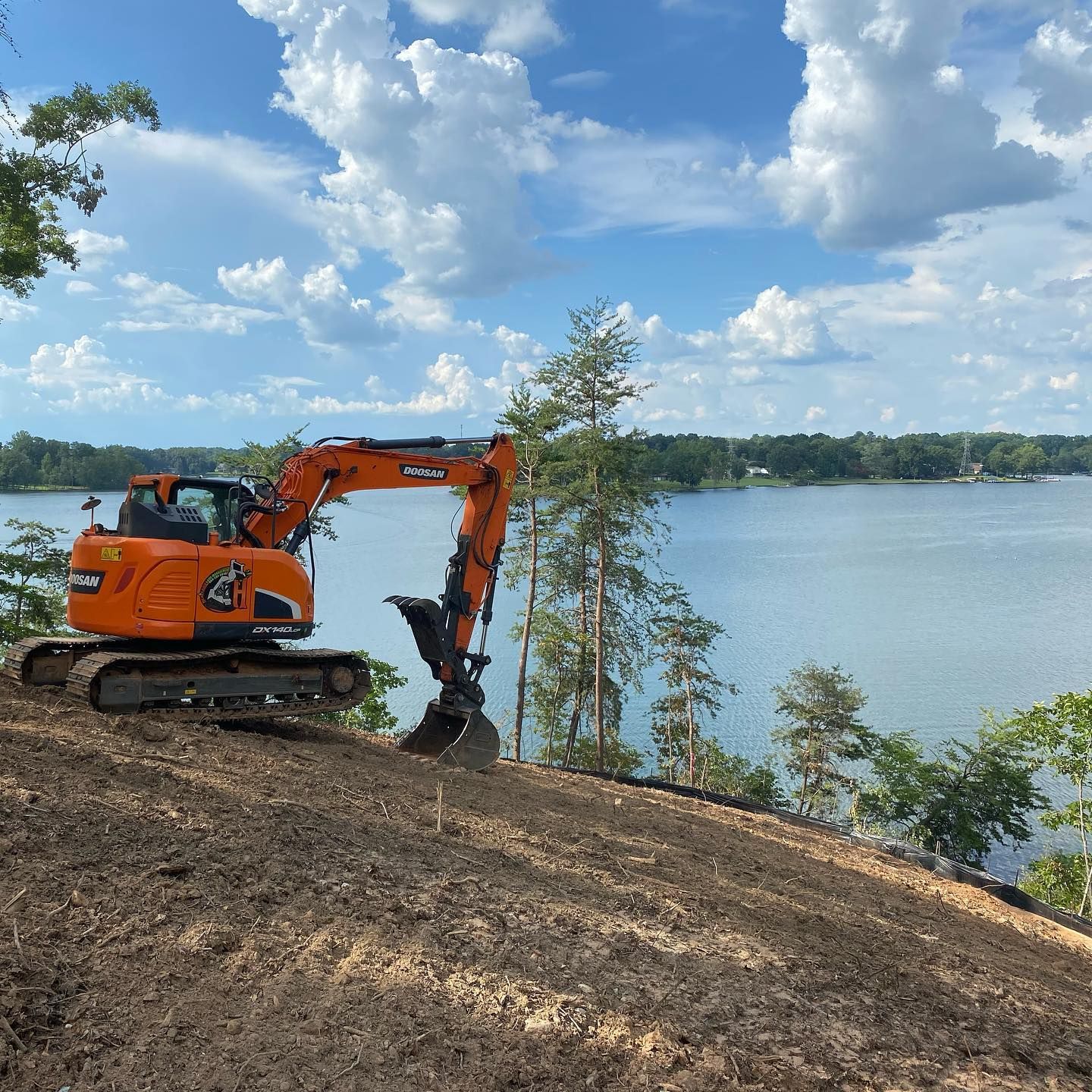 Orange excavator on a hillside by a lake under a cloudy blue sky, actively digging.