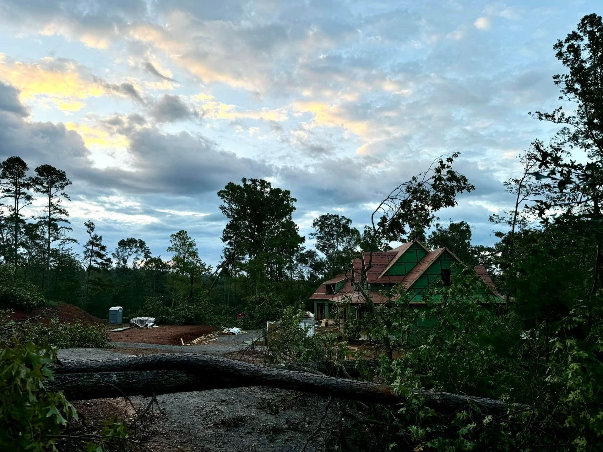 Sunset sky over a partially built house surrounded by trees; a fallen log is in the foreground.