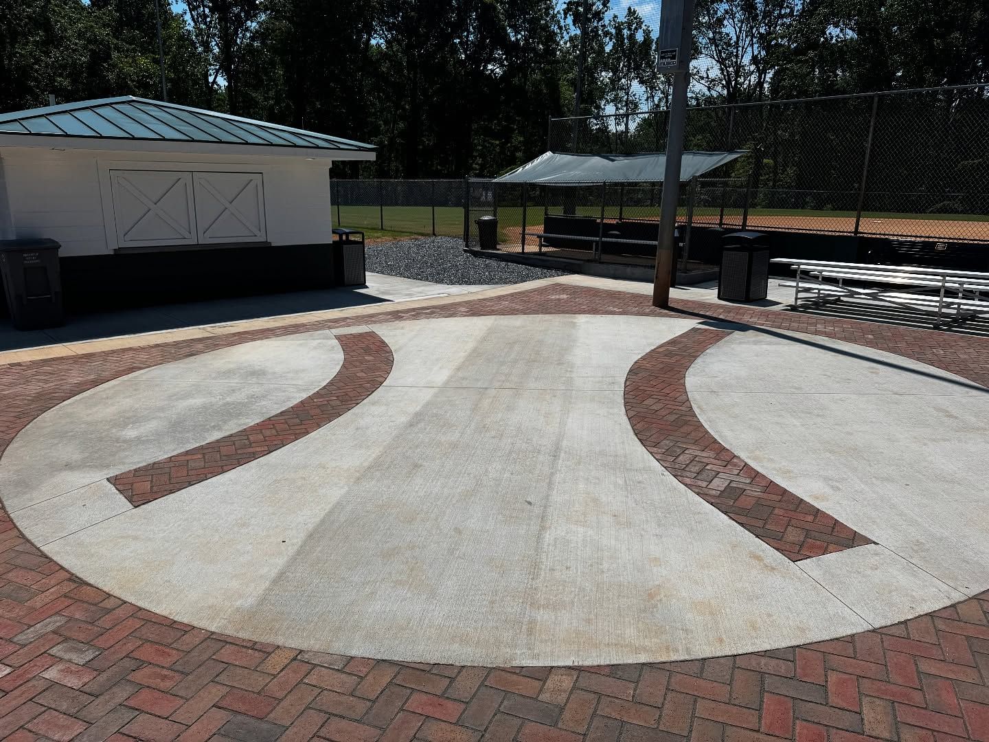 Brick and concrete patio with curved red brick accents, near a shelter and baseball field.
