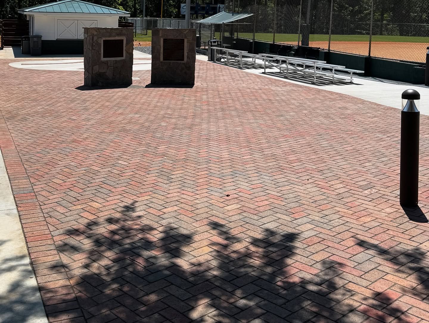 Brick-paved walkway with two stone pillars, a baseball field, and tree shadows.