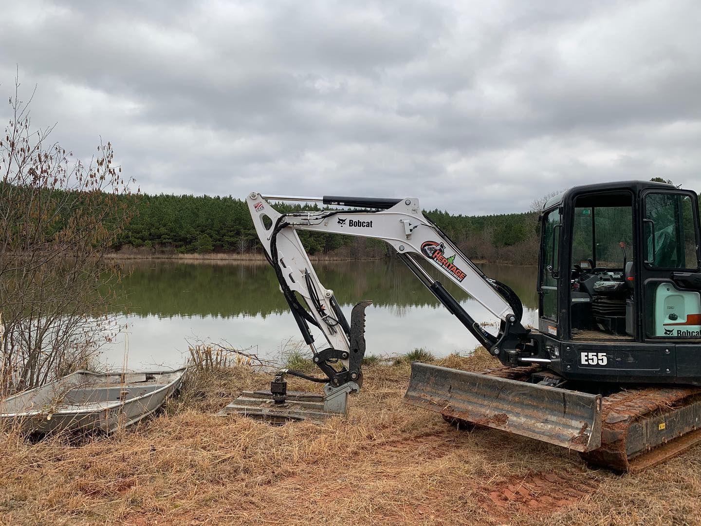 Small excavator at edge of a lake, next to a boat. Overcast sky, trees in the background.