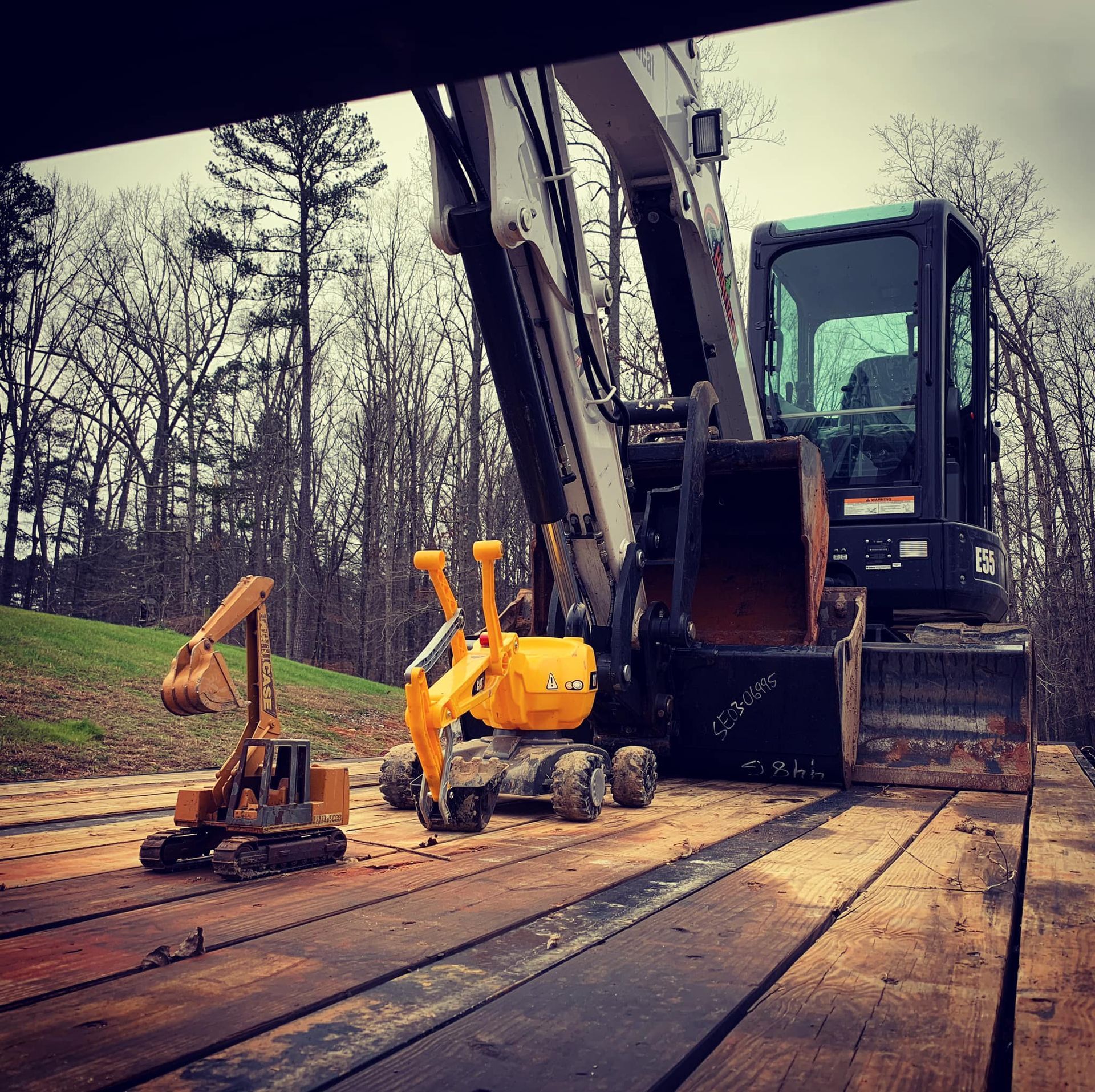 Excavators on a trailer, one large, two small, in a wooded area with bare trees.