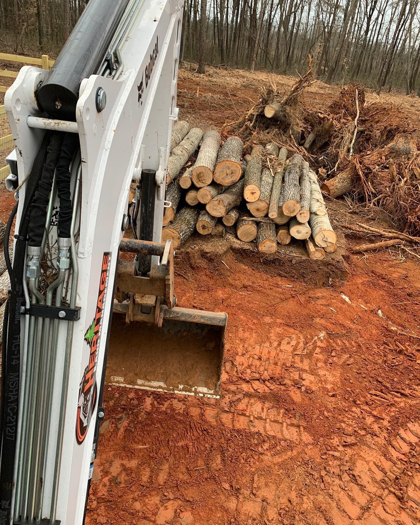 Bobcat excavator with a metal bucket, red dirt ground, and a pile of logs.