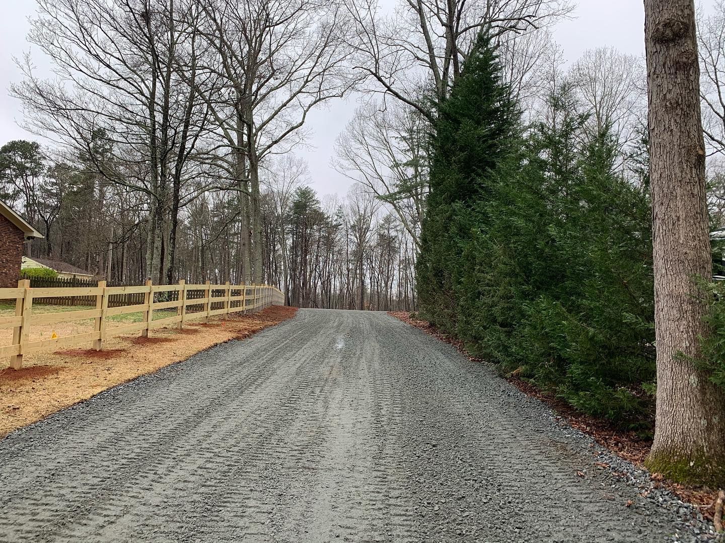 Gravel driveway leading into a wooded area, bordered by a split rail fence and evergreen trees under a cloudy sky.