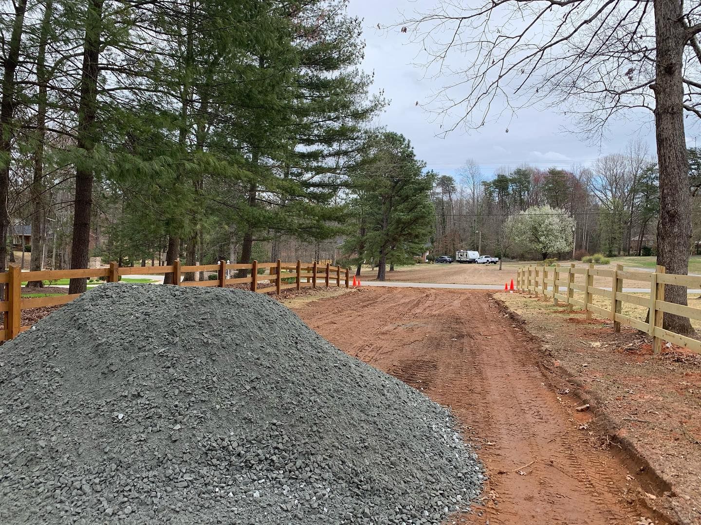 Pile of gravel on a dirt driveway leading to a wooden fence and trees on an overcast day.