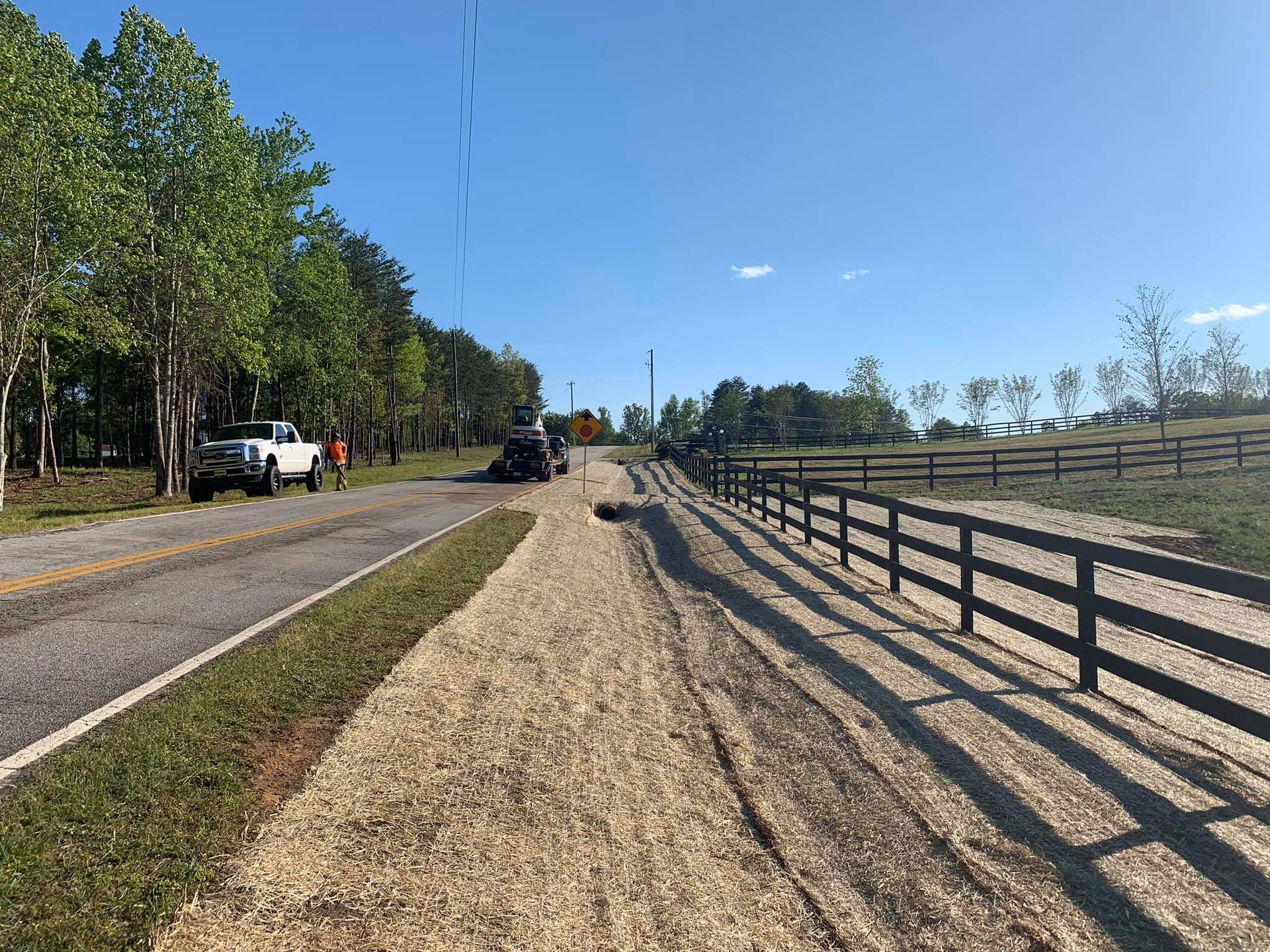 A gravel driveway next to a road, with a fence on the right, trees and a truck. Blue sky.
