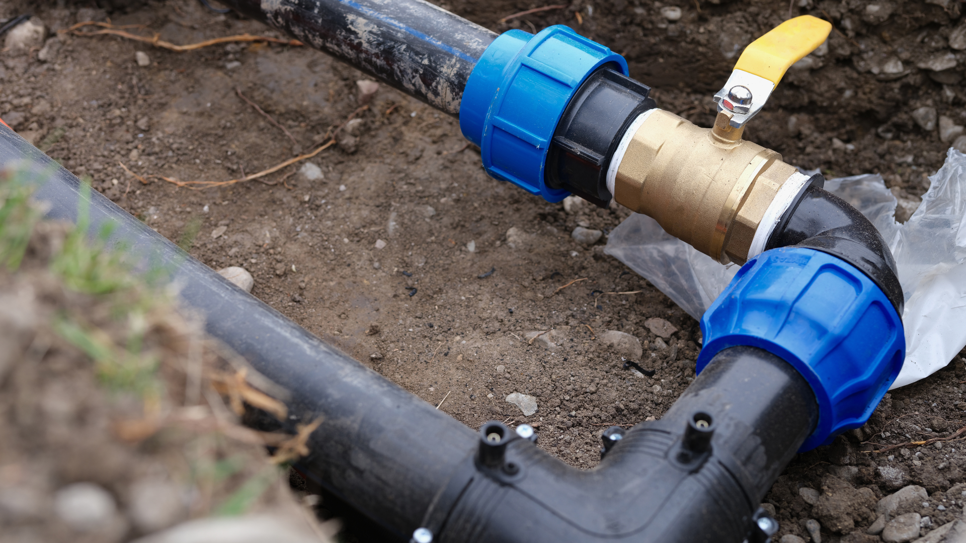 Black PVC pipes and blue couplings connected to a brass valve in a dirt trench.