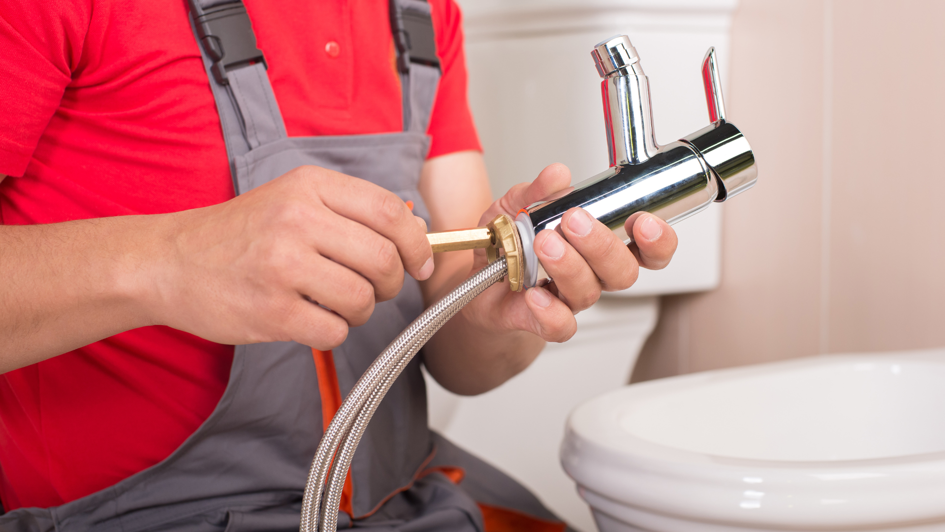 Plumber installing faucet in bathroom.  Silver faucet, braided hose, red shirt, gray overalls.