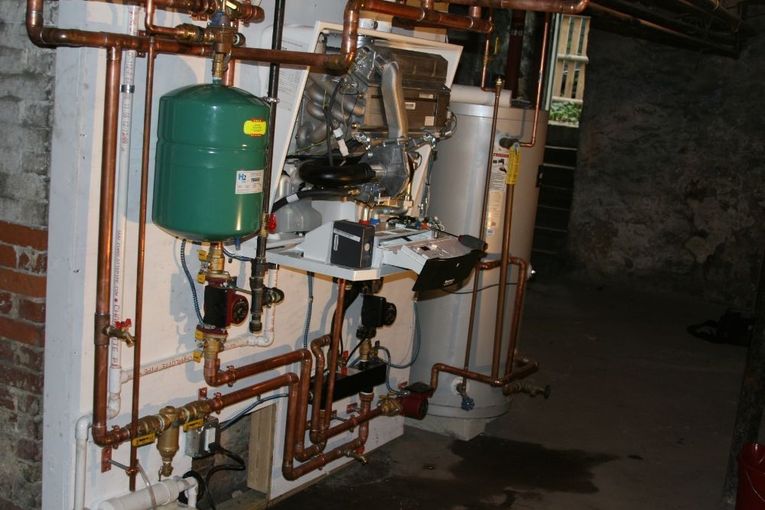 Complex plumbing system with copper pipes and green tank mounted on a white wall in a basement.
