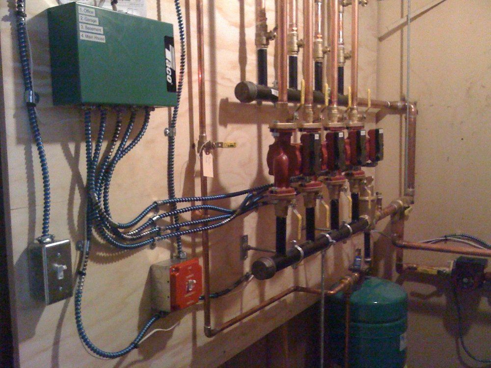 Hydronic heating system with copper pipes, valves, and green electrical box on plywood wall.