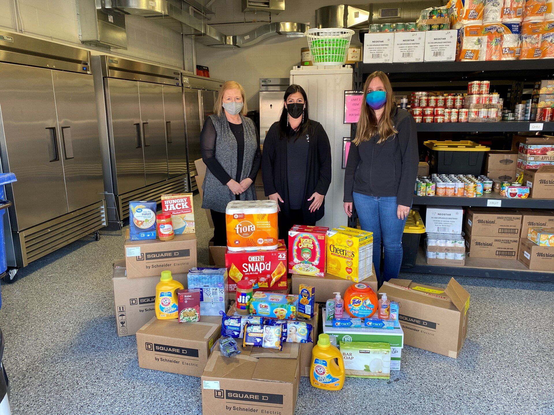 Three women wearing masks are standing in a room filled with boxes of food.