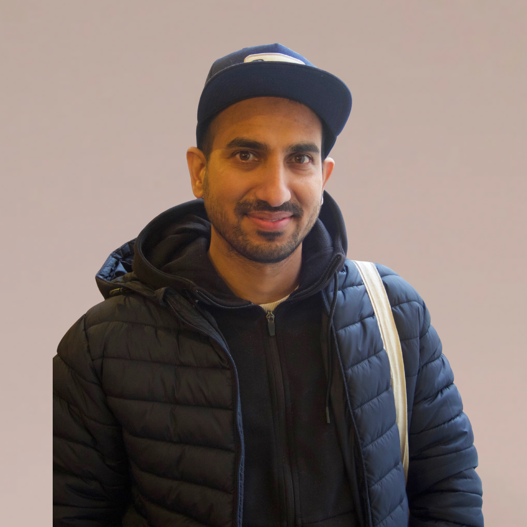 Mann i mørk jakke og baseballcaps smiler, skulderveske, nøytral bakgrunn.