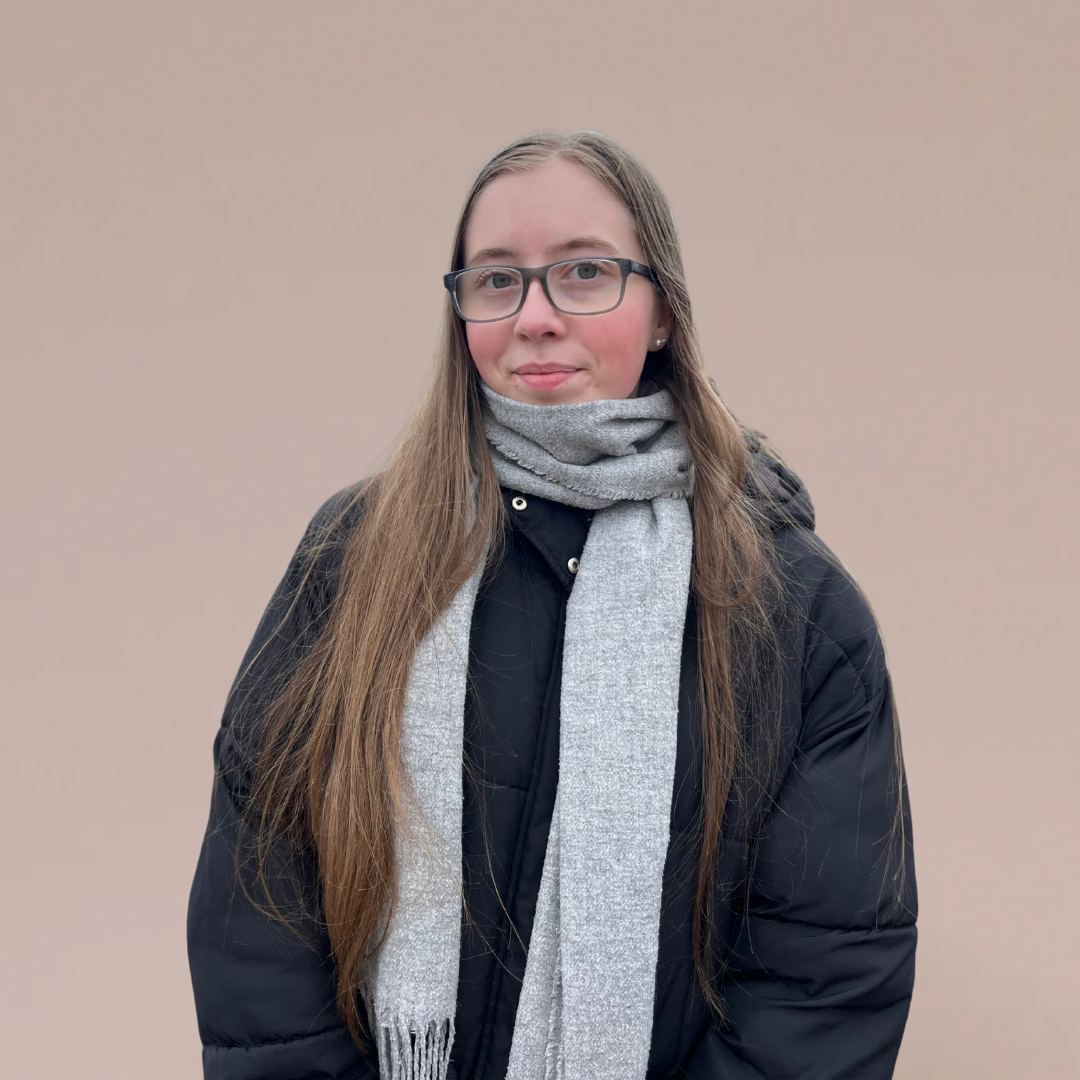 Kvinne med langt brunt hår, briller og grått skjerf foran en enkel rosa bakgrunn.