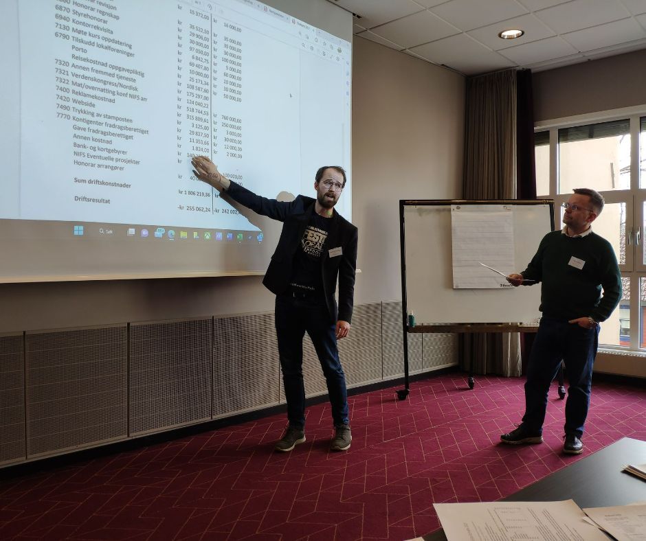 To menn er til stede på en konferanse, den ene peker på et regneark på en skjerm, den andre holder en penn, i et rom med rød løper.